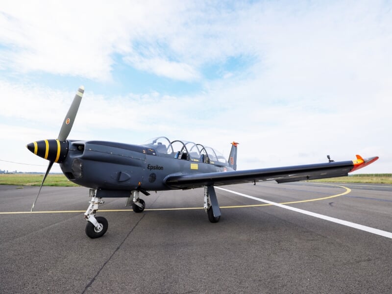 Avion d'entraînement Epsilon gris stationné sur une piste d'aéroport sous un ciel partiellement nuageux