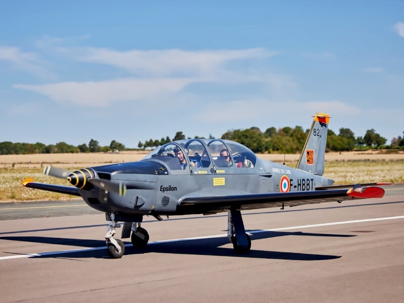 Petit avion militaire gris avec deux pilotes à bord, prêt au décollage sur une piste d'aéroport par temps clair.