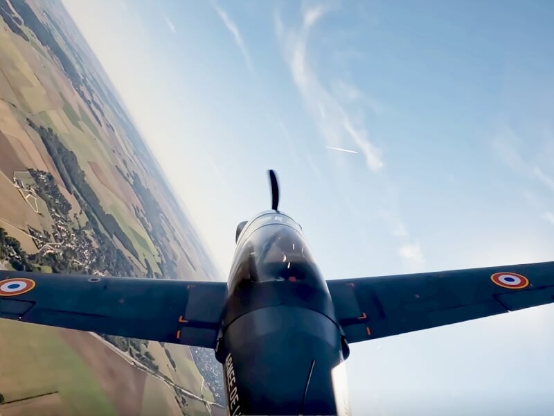 Vue en vol d'un avion de voltige militaire avec ses ailes déployées au-dessus d'un paysage rural et ciel bleu clair.
