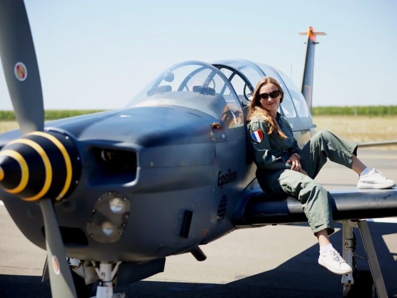 Jeune femme en combinaison de pilote assise sur l'aile d'un avion de voltige bleu sur piste d'aéroport ensoleillée.