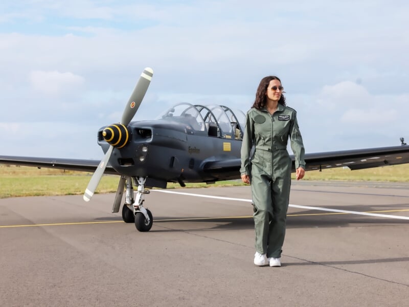 Femme pilote en combinaison verte marchant sur la piste devant un avion d'entraînement gris à hélice au sol