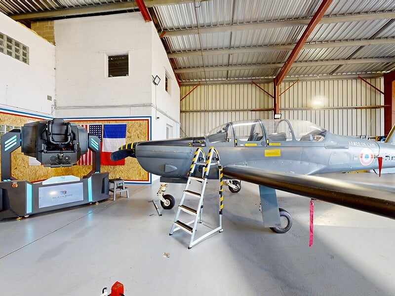 Vue d’ensemble d’un hangar avec un avion d’entraînement militaire à côté d’un simulateur de vol gyroscopique.