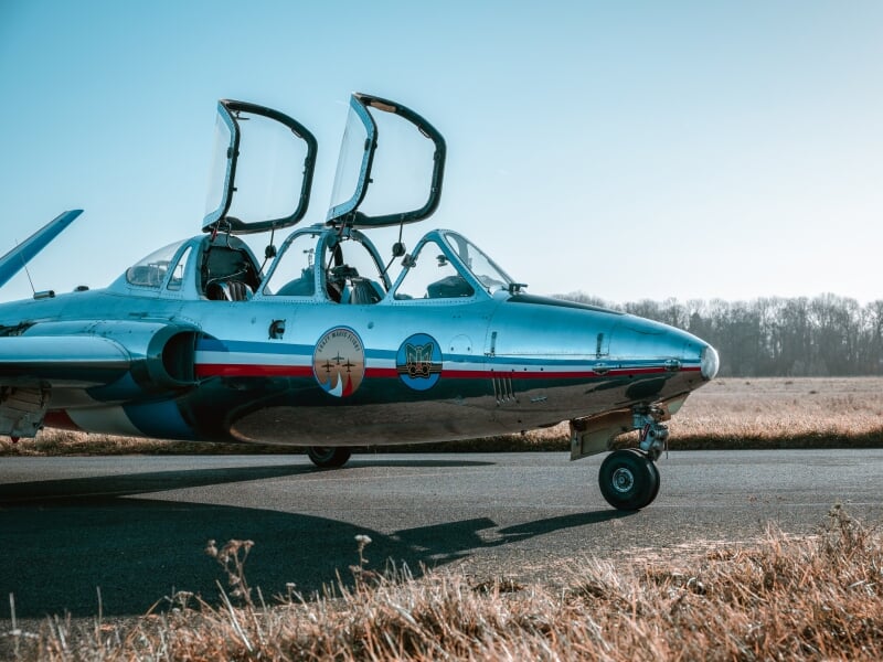 Alpha Jet stationné sur une piste, coques de cockpit ouvertes, décoré de deux insignes et bandes tricolores, ciel bleu en arrière-plan.