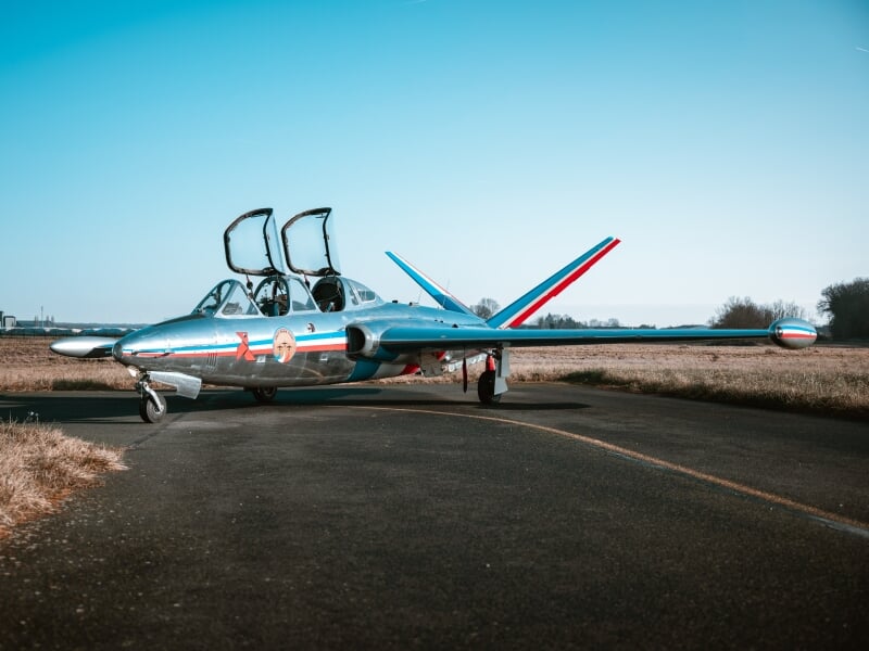 Avion de chasse argenté stationné sur une piste d'aéroport avec les cockpits ouverts sous un ciel bleu clair.