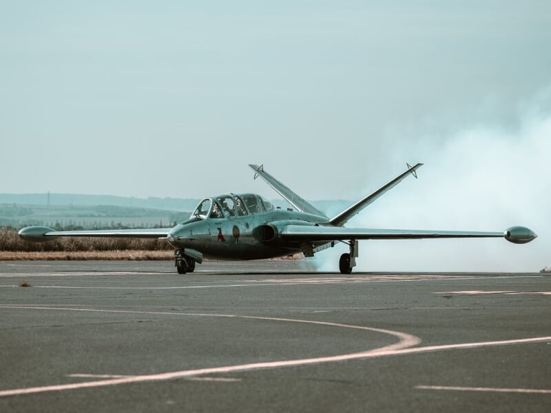 Alpha Jet au sol, émettant de la fumée, sur une piste avec arrière-plan champêtre et ciel nuageux.