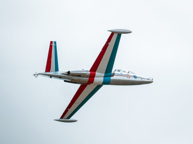 Alpha Jet en vol, vu de profil, décoré de bandes rouge et bleue sur fond de ciel gris clair.