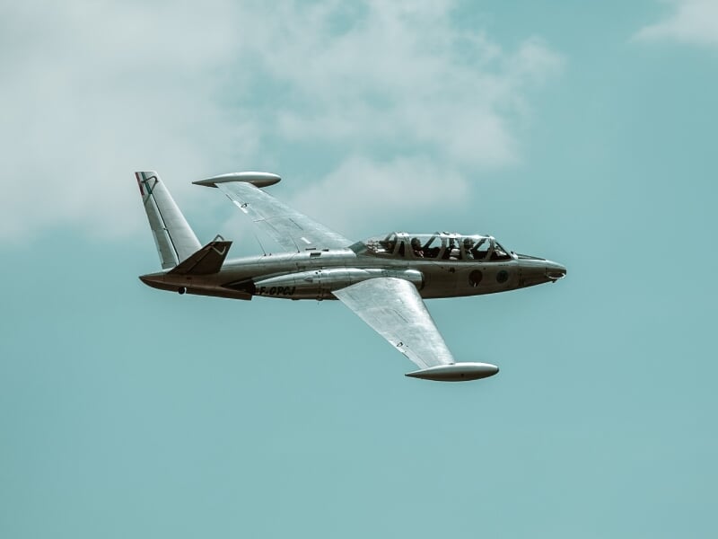 Alpha Jet en vol sous un ciel bleu, vue latérale, avec deux pilotes à bord, ailes et fuselage argentés.