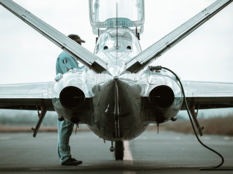 Technicien en combinaison bleu clair effectuant des vérifications sur un Alpha Jet au sol, vue arrière avec deux réacteurs visibles.