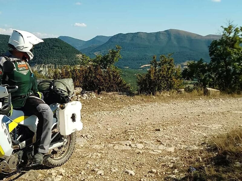 Motard en tenue complète sur sa moto tout-terrain sur un chemin rocailleux avec montagnes en arrière-plan.