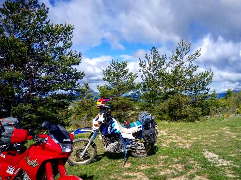 Deux motos garées sur une prairie verte avec des arbres et un ciel partiellement nuageux en arrière-plan.