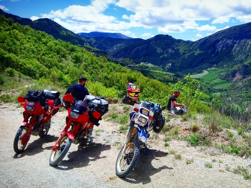 Quatre motos et trois motards sur un chemin en hauteur avec une vue panoramique sur une vallée verdoyante.