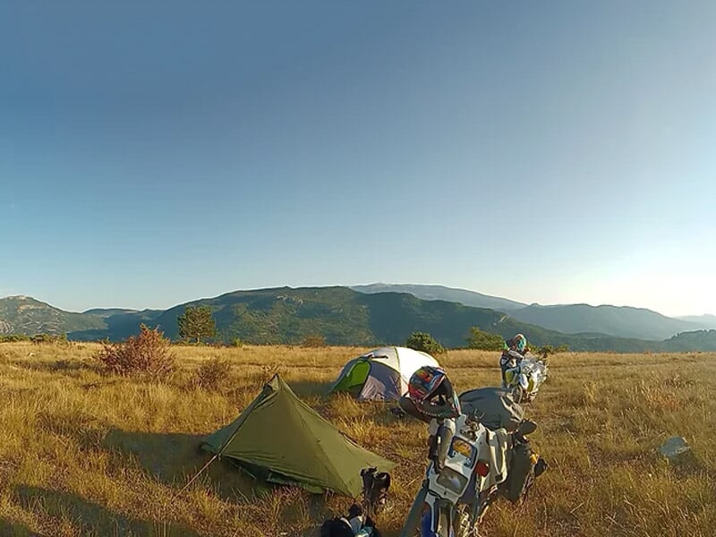Deux motos stationnées près de tentes dans un champ sec avec des collines et un ciel clair au coucher du soleil.