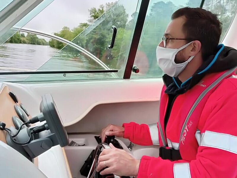 Homme portant un masque et un gilet de sauvetage rouge, conduisant un bateau sur une rivière calme sous un ciel nuageux.