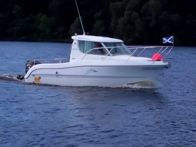 Bateau blanc naviguant sur un lac ou une rivière, avec un drapeau écossais à l'arrière et une forêt dense en arrière-plan.