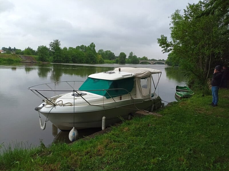 Petit bateau amarré au bord d'une rivière, entouré d'herbe verte et d'arbres, avec une personne debout à proximité.