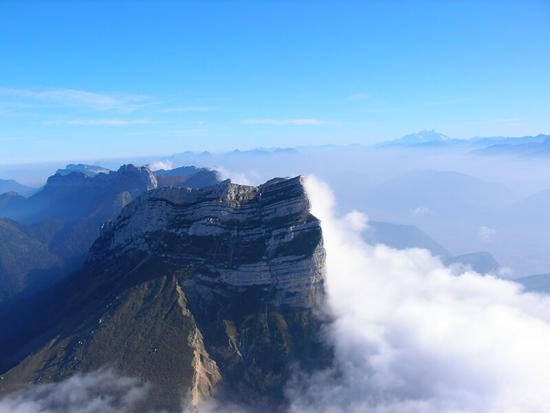 Vue aérienne d'une montagne escarpée avec des nuages blancs flottant autour des sommets sous un ciel bleu clair.