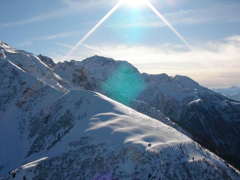 Paysage de montagnes enneigées sous un ciel bleu avec un soleil brillant et un léger halo lumineux.