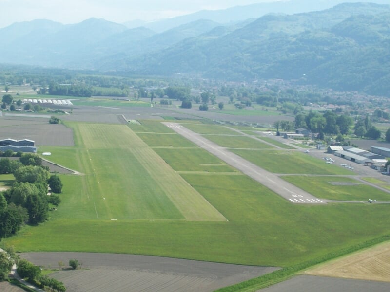Vue aérienne d'un aérodrome avec une piste d'atterrissage entourée de bâtiments et de collines verdoyantes.