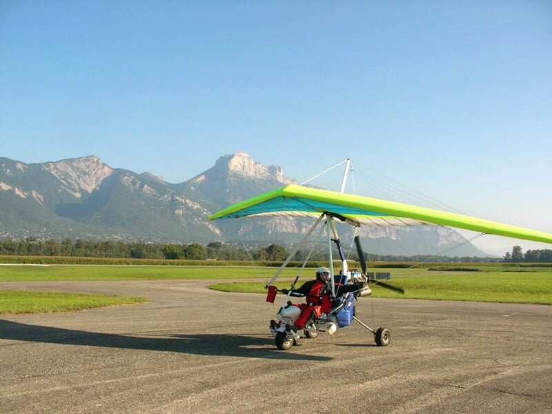 ULM avec une aile verte posé sur une piste d'atterrissage, avec des montagnes en arrière-plan sous un ciel bleu.