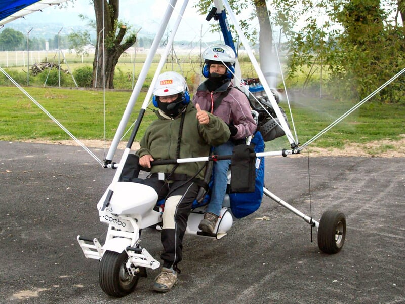 Deux personnes équipées de casques sont assises dans un ULM tricycle sur une piste goudronnée entourée d'herbe verte.