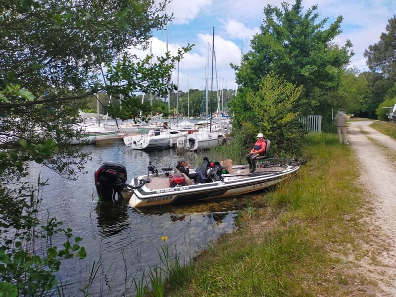 Bateau de pêche amarré au bord d'un canal avec des voiliers en arrière-plan lors d'un séjour avec un guide de pêche sur les lacs des Landes.