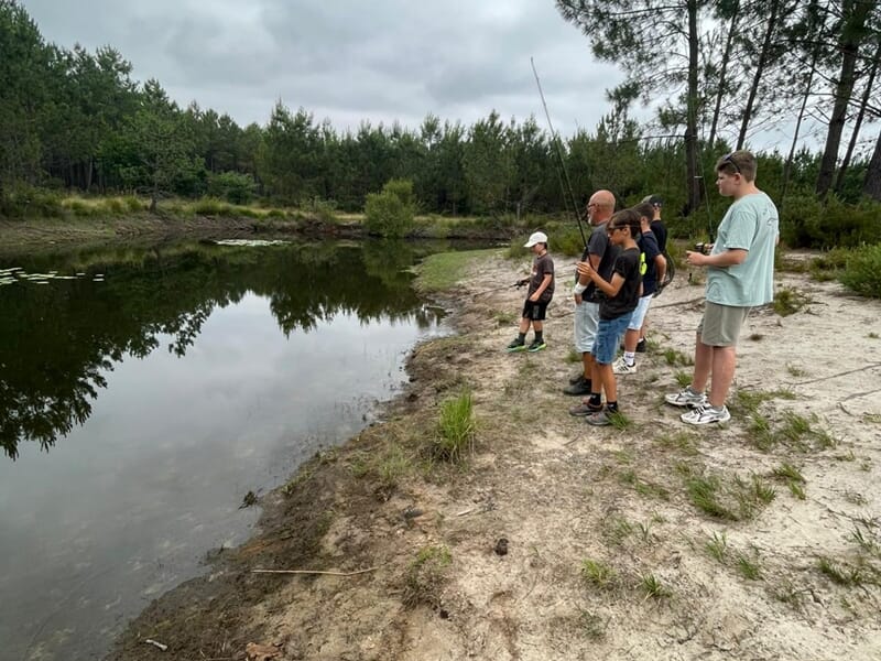 Groupe de personnes pêchant au bord d'un plan d'eau entouré de pins lors d'un stage de pêche pour enfant sur les lacs des Landes.