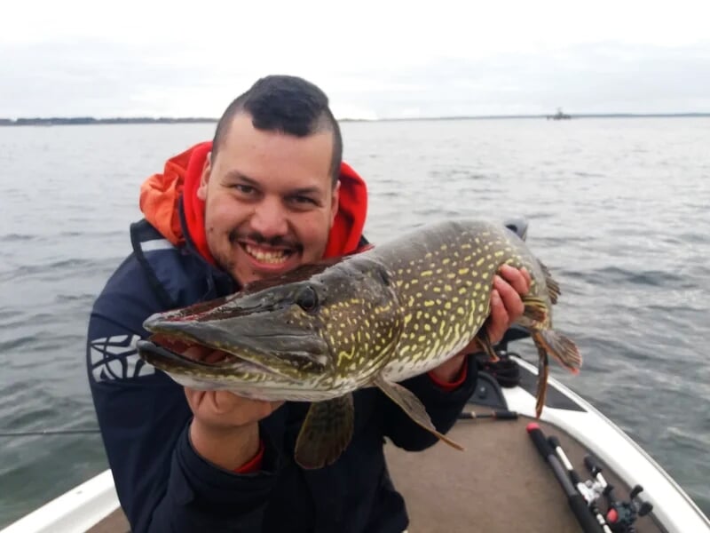 Homme souriant tenant un grand brochet fraîchement pêché sur un bateau au milieu d'un lac calme sous un ciel gris.