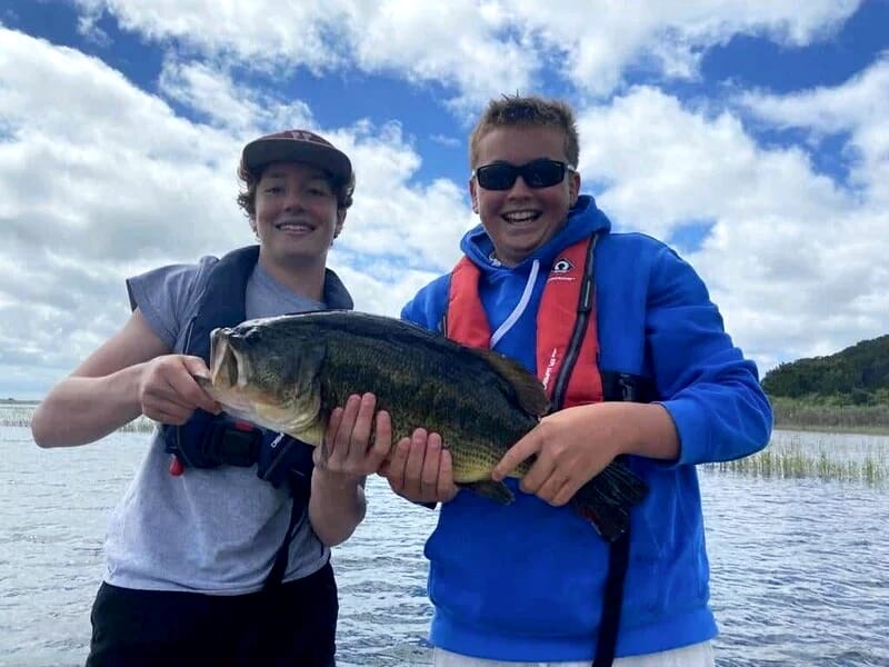 Deux jeunes pêcheurs heureux portant des gilets de sauvetage, tenant un gros poisson noir et vert sur un lac avec ciel nuageux.
