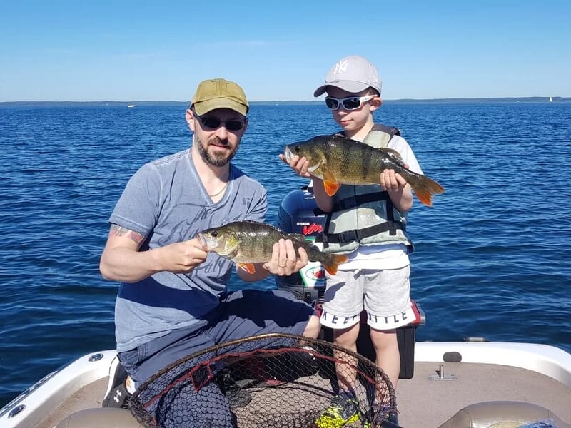 Un homme et un enfant sur un bateau, chacun tenant un poisson, avec un filet de pêche posé devant eux sur un lac bleu clair.