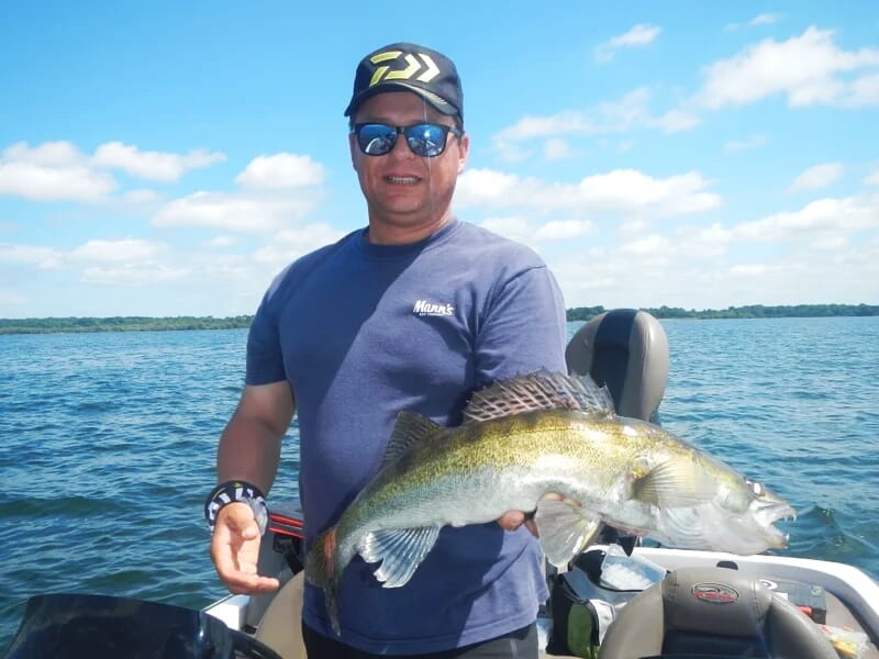 Homme en lunettes de soleil et casquette tenant un gros poisson jaune-vert sur un bateau, avec un ciel bleu et quelques nuages.