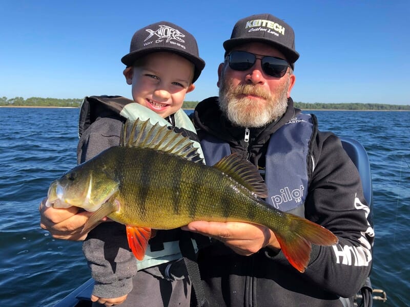 Un homme et un enfant souriants sur un bateau, tenant un gros poisson jaune et noir fraîchement pêché.