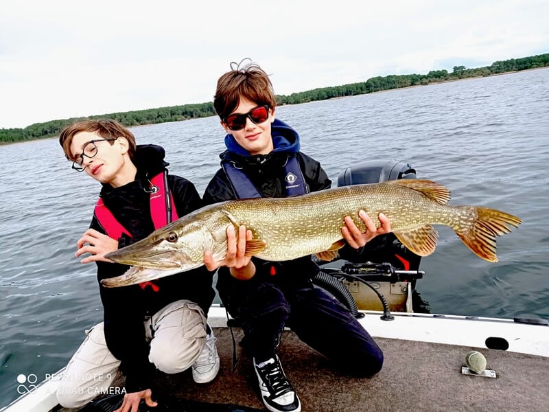 Deux garçons assis dans un bateau, tenant un grand brochet capturé lors d'une sortie de pêche en lac.