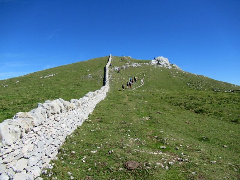 Sentier de randonnée montant une colline verte bordée d'un mur en pierres sèches sous un ciel bleu
