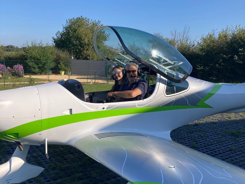 Deux personnes assises dans un avion ultraléger blanc et vert avec un cockpit ouvert sous un ciel bleu clair.