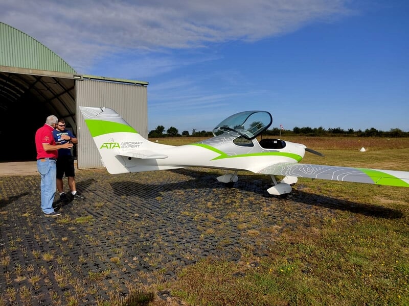 Un avion ultraléger blanc et vert stationné devant un hangar avec deux hommes discutant à côté.