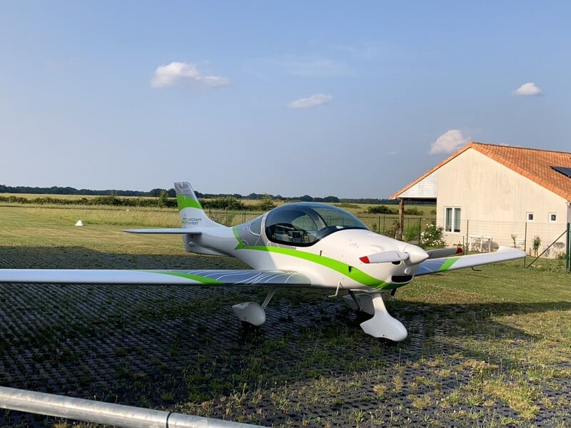 Avion ultraléger blanc et vert posé sur une piste avec une maison en arrière-plan sous un ciel partiellement nuageux.