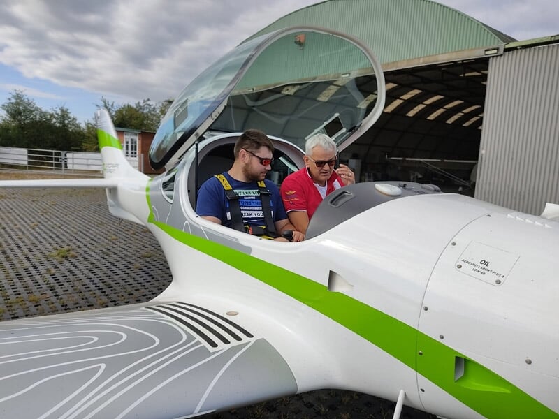 Deux hommes assis dans un avion ultraléger blanc et vert avec le cockpit ouvert devant un hangar.