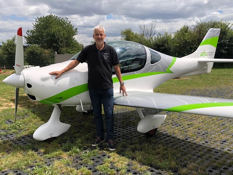 Homme debout souriant appuyé sur l'aile d'un avion ultraléger blanc et vert sur une piste herbeuse.