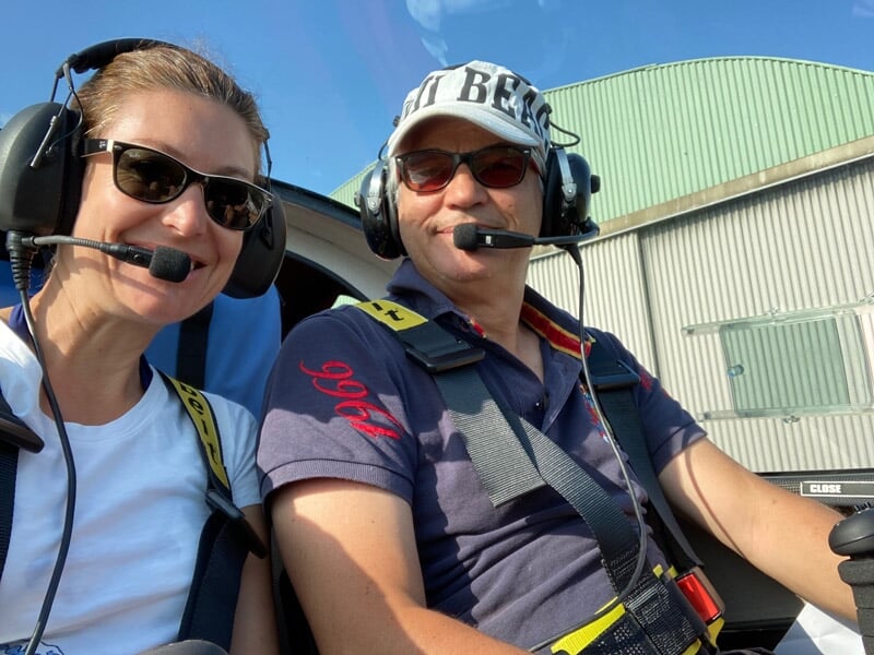 Deux personnes souriantes portant des casques de pilote assises dans un avion ultraléger blanc et vert.