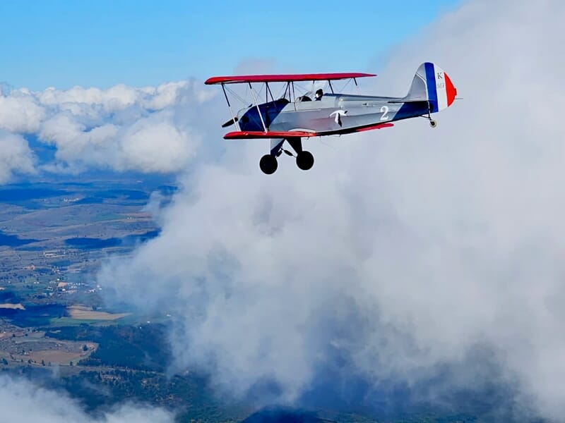 Petit avion biplan rouge et blanc volant au-dessus des nuages avec un paysage rural en dessous.