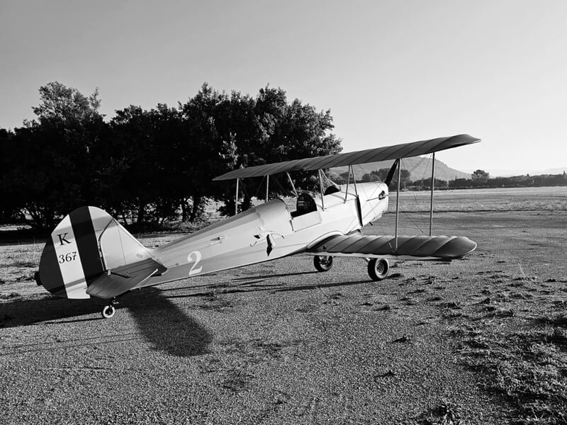 Avion biplan immobile sur une piste, photo en noir et blanc, avec un arbre en arrière-plan.