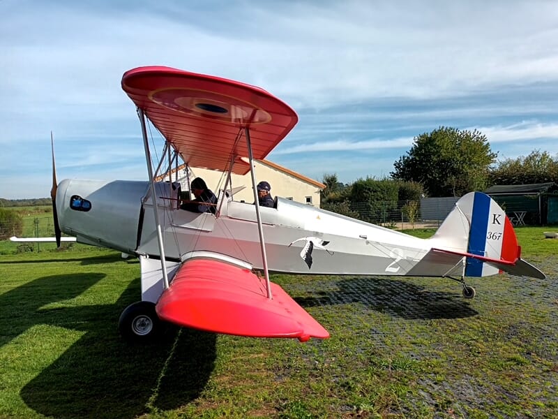 Avion biplan rouge et blanc stationné sur une pelouse verte avec deux personnes à bord.