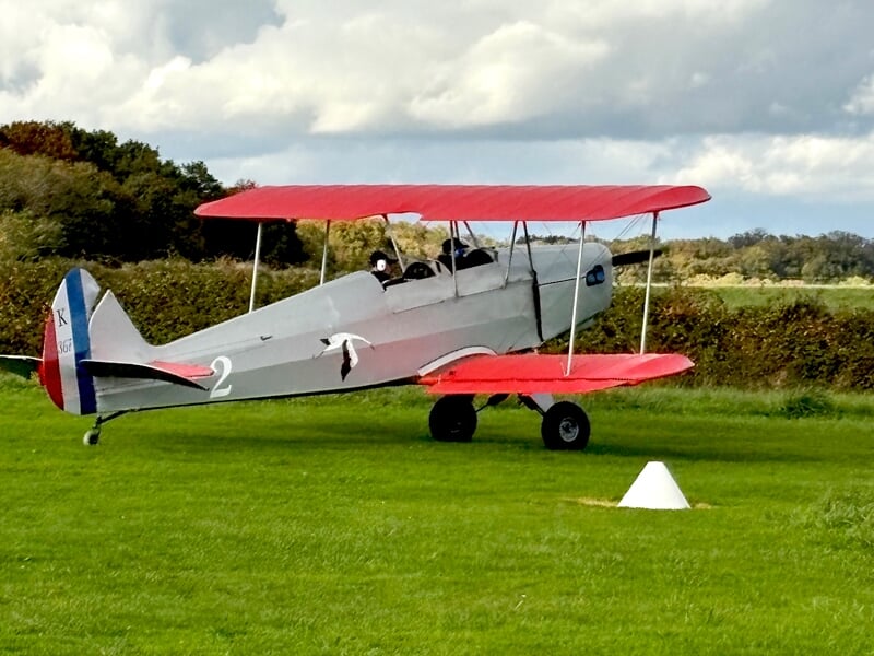 Avion biplan rouge et blanc sur une pelouse verte, prêt au décollage avec un ciel nuageux.