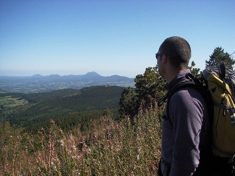 Homme avec sac à dos regardant un vaste paysage de collines et forêts sous un ciel bleu.