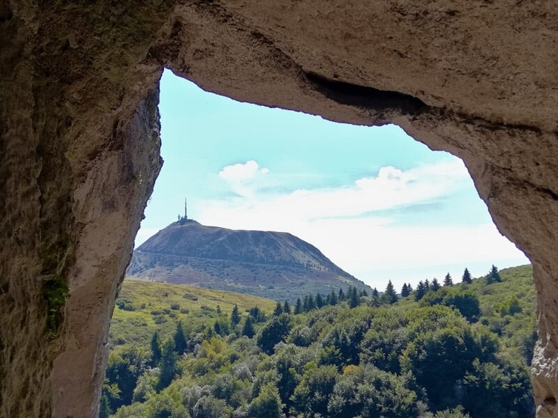 Vue encadrée par une ouverture rocheuse sur une montagne avec un sommet marqué par une antenne.