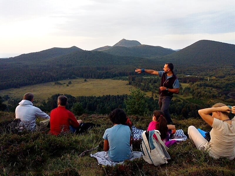 Groupe de personnes assises sur une colline, écoutant un guide qui montre le paysage volcanique.