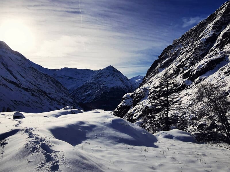 Paysage de vallée enneigée avec traces de pas et montagnes en arrière-plan sous un ciel clair.
