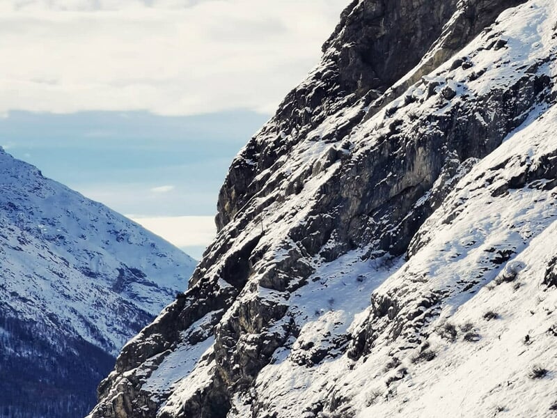 Vue rapprochée d'une paroi rocheuse enneigée avec montagnes en arrière-plan sous un ciel bleu.