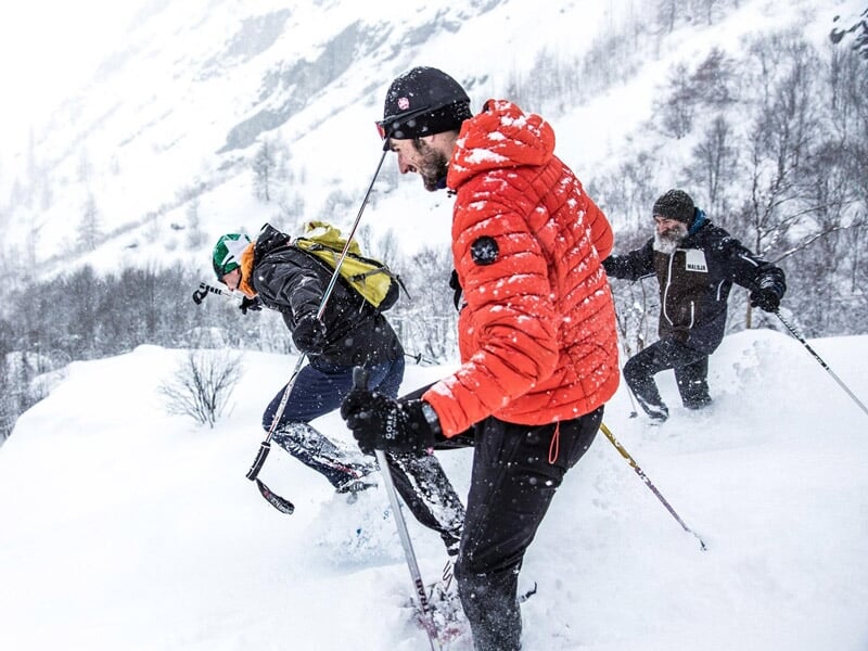 Trois randonneurs en raquettes à neige dans un paysage blanc avec arbres et montagnes en arrière-plan.