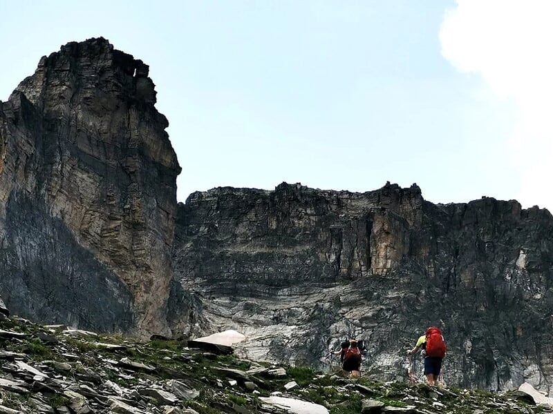 Deux randonneurs avec sacs à dos marchant sur un sentier rocheux en montagne sous un ciel bleu.